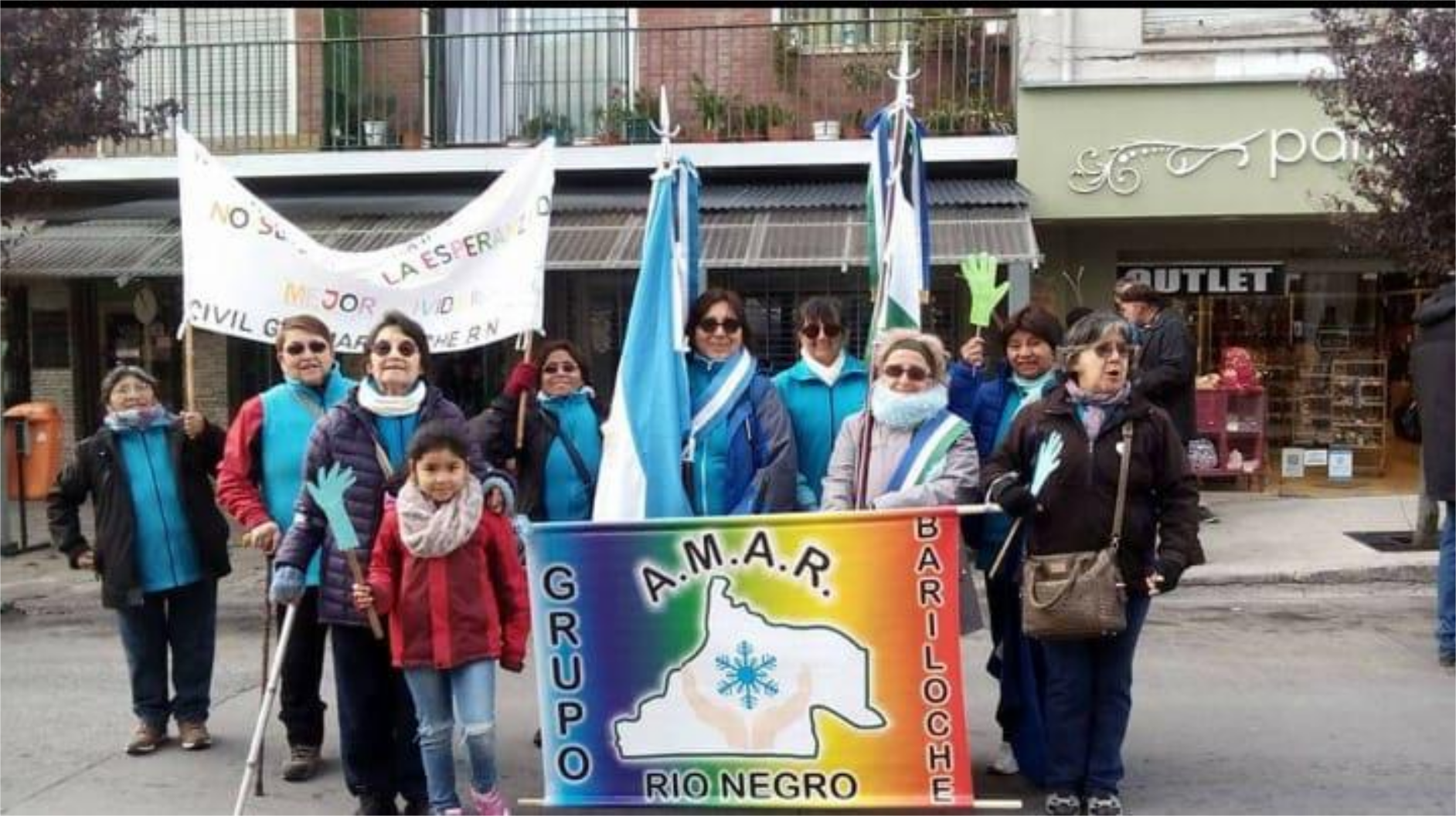 Fotografía de varias personas en la calle durante una actividad. Una mujer lleva la bandera y la banda de Argentina, y a su lado, otra mujer lleva la bandera y la banda de Río Negro. Además, frente a ellas sostienen un banner de "Grupo Amar"
